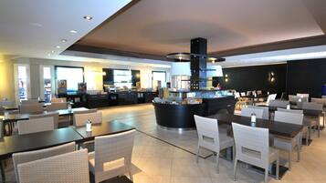 A modern restaurant with a central bar, multiple dining tables, and wicker chairs.