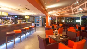 A modern restaurant with orange and grey seating, a bar area with a variety of bottles, and a large window offering a view of the city. - Pascal Paoli (Aregno)