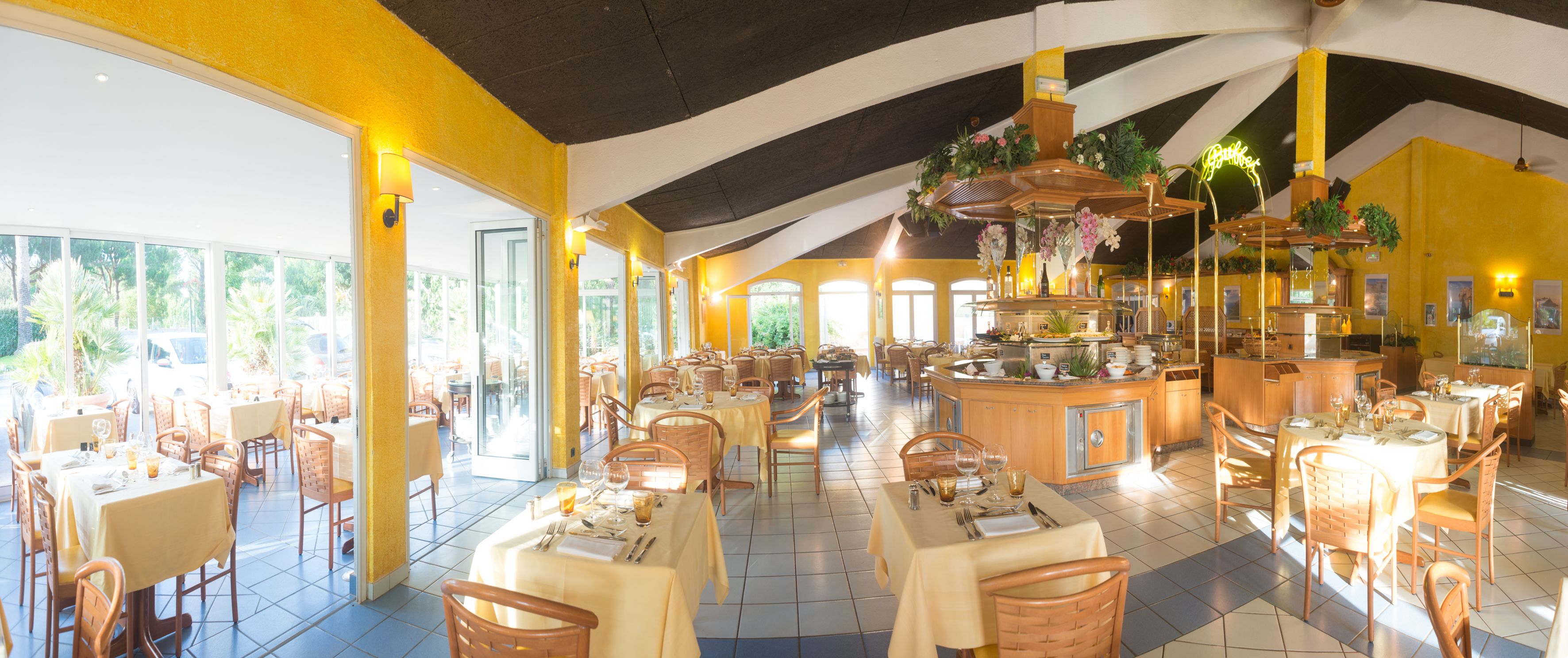 Un restaurante con paredes amarillas, ventanas amplias y mobiliario de madera. Las mesas están dispuestas con manteles blancos y hay plantas colgantes sobre la zona del bar.