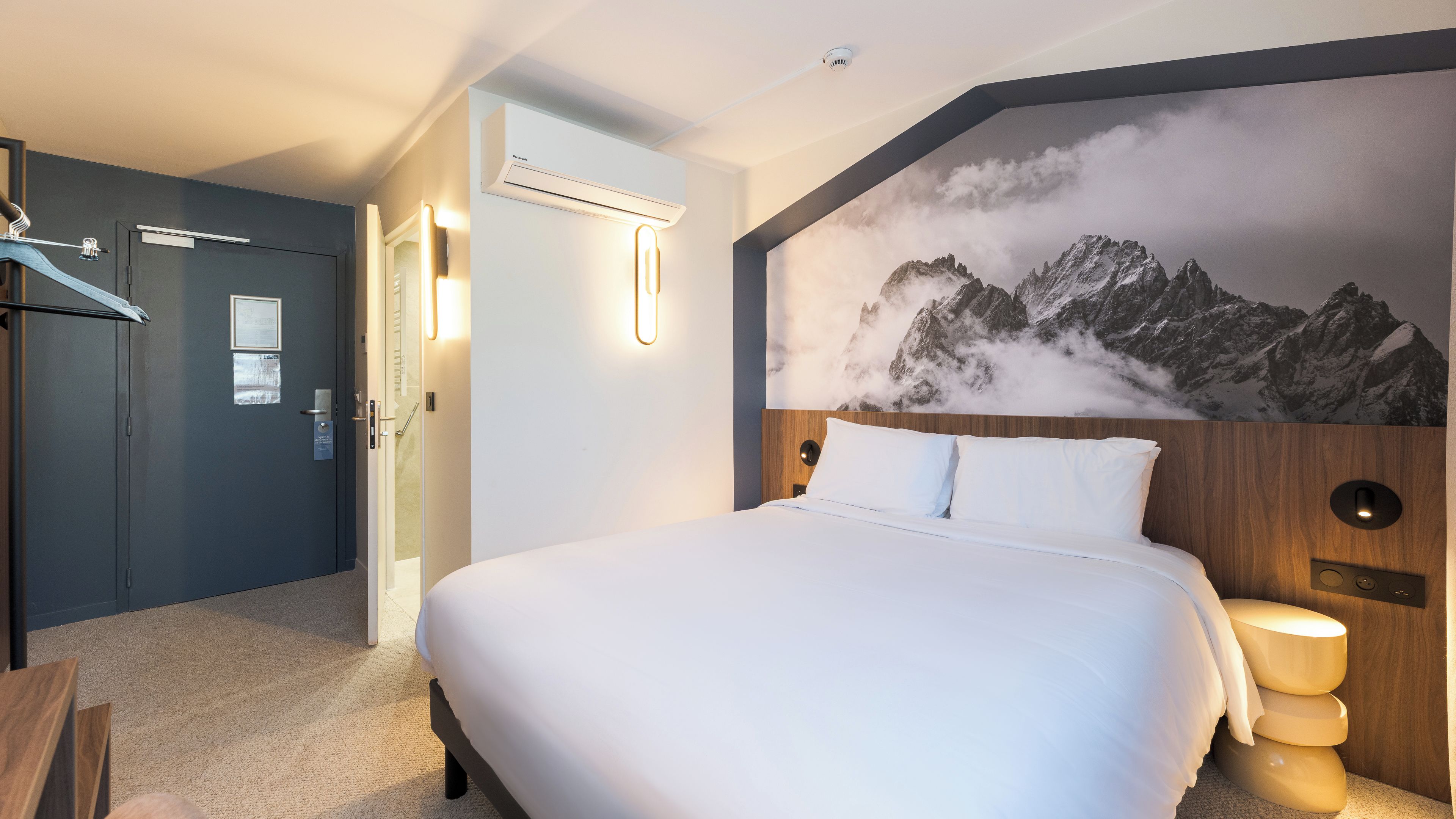 A hotel room with a large bed, a wooden headboard, a wall-mounted air conditioner, and a mountain mural.