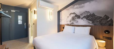 Una habitación de hotel con una cama grande, cabecera de madera, aire acondicionado de pared y un mural de montañas.
