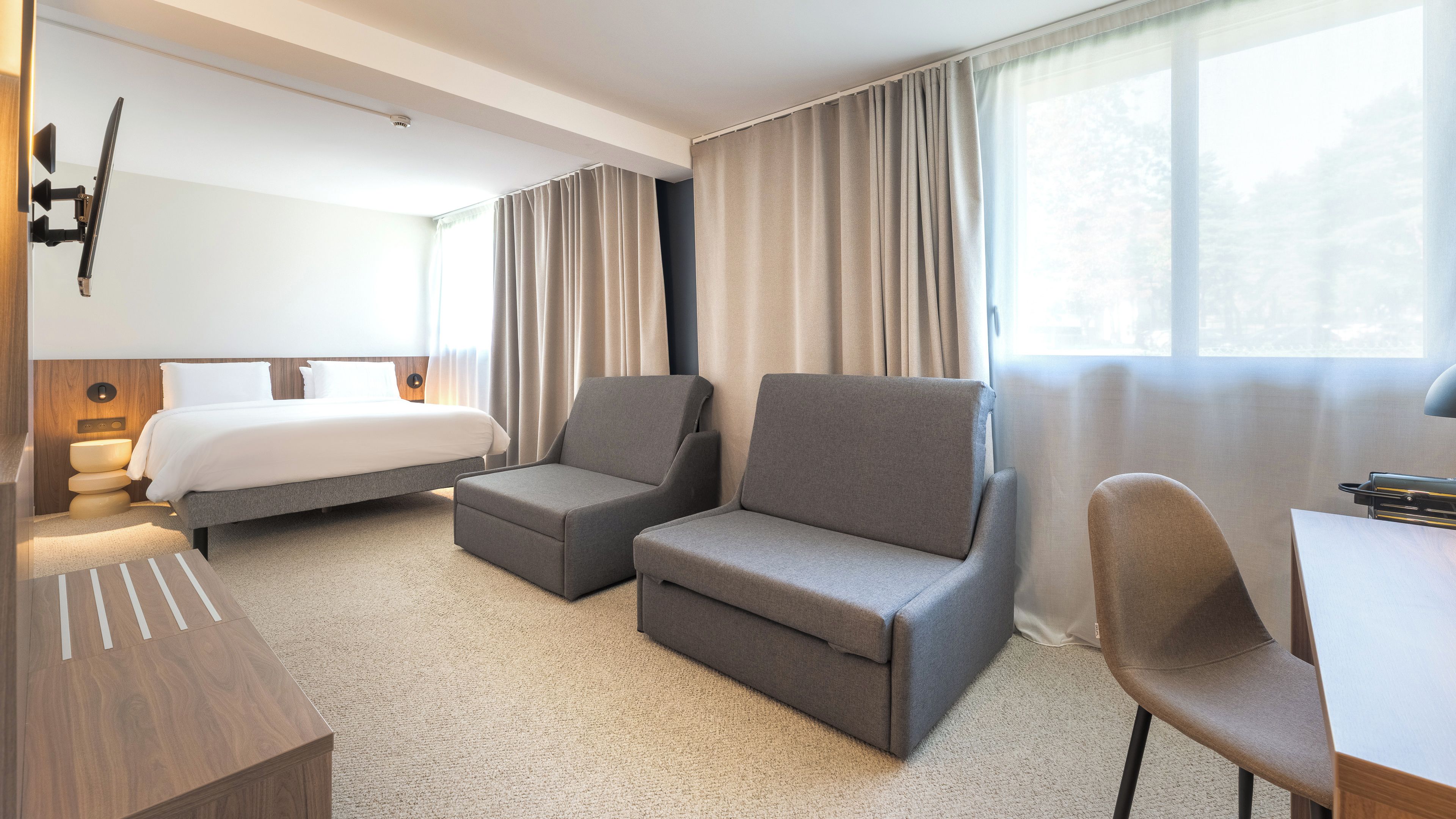 A hotel room with a bed, two armchairs, a desk, and a window with curtains.