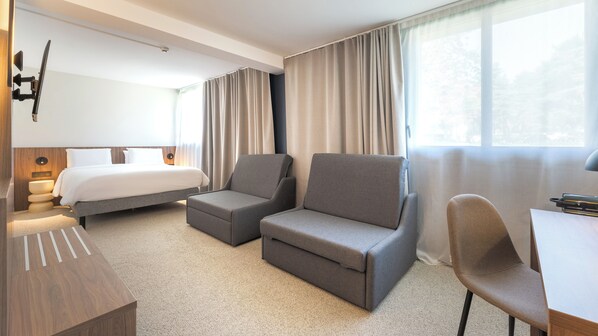 Une chambre d’hôtel avec un lit, deux fauteuils, un bureau et une fenêtre avec des rideaux.