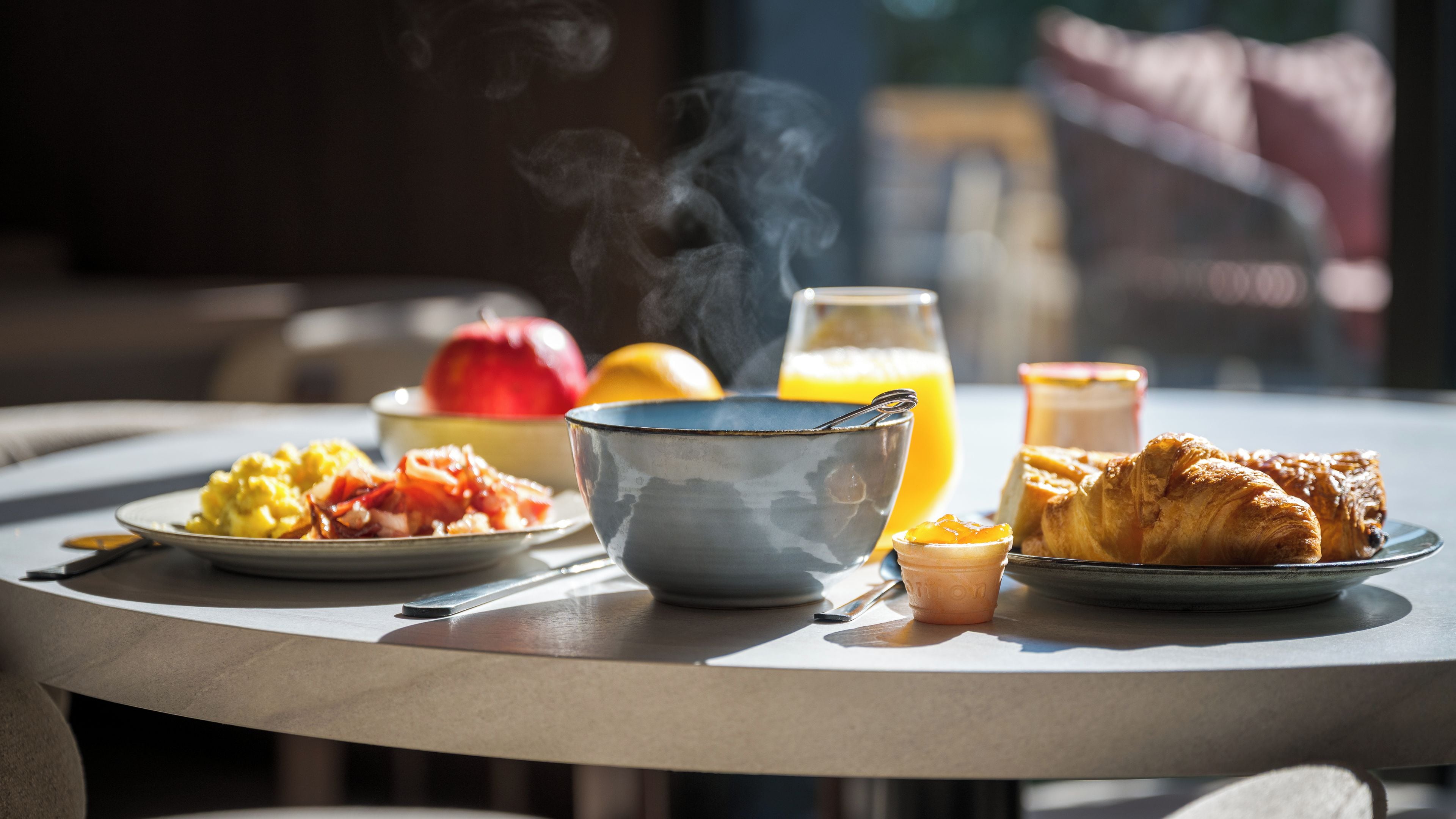 A breakfast table with a bowl, a plate of food, a croissant, and a glass of orange juice.