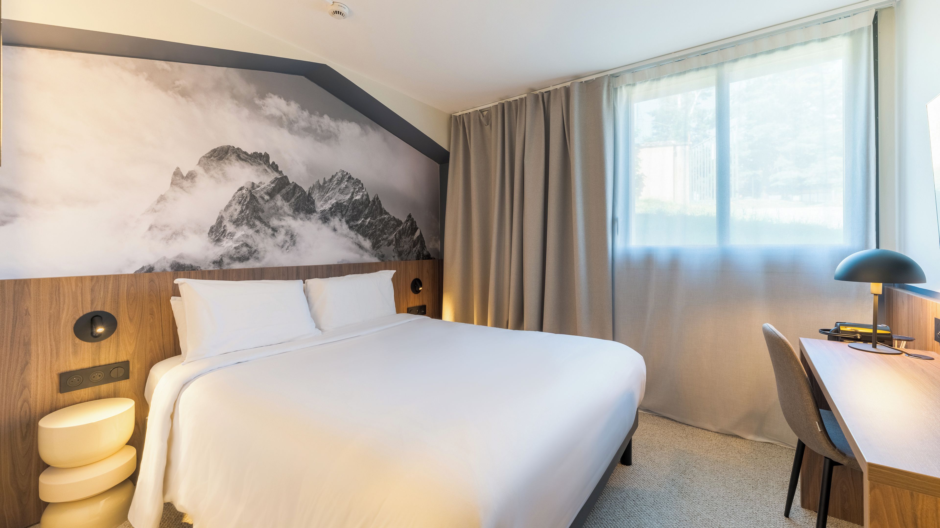 Een hotelkamer met een bed, een bureau, een stoel, een wandlamp en een raam met gordijnen.
