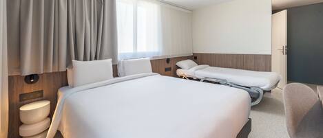 A hotel room with a large bed, two smaller beds, a bedside table, a chair, a toilet, and a window with curtains.