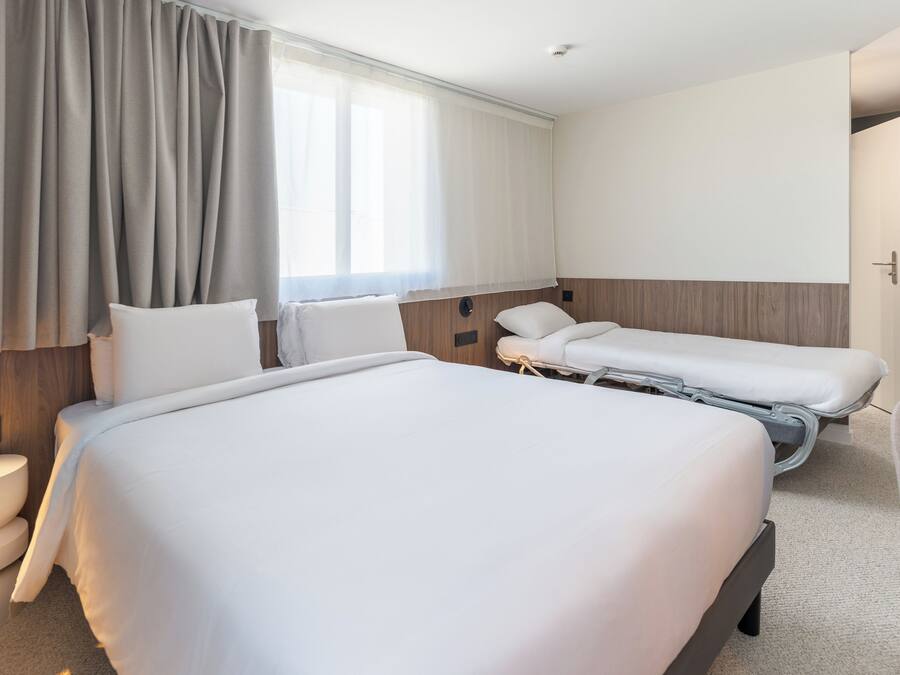 Hotellihuone, jossa on suuri sänky, kaksi pienempää sänkyä, yöpöytä, tuoli, wc ja ikkuna, jossa on verhot.