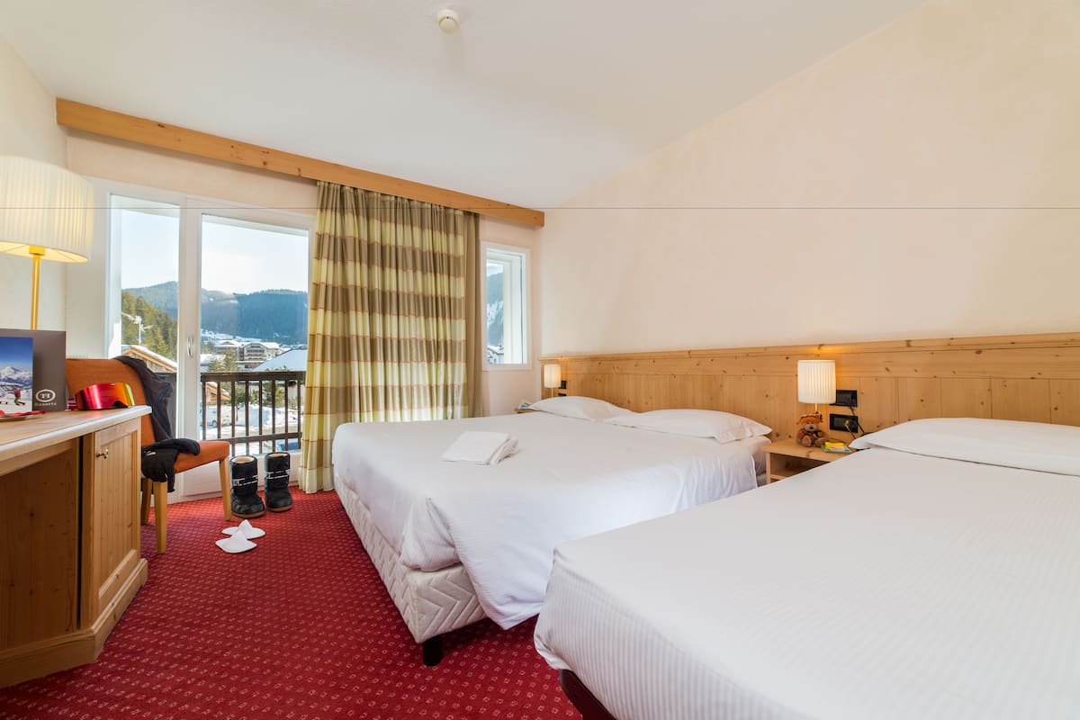 A hotel room with two beds, a desk with a chair, a small table, and a window with a view of a bridge and mountains.
