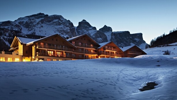A large lodge with multiple wooden buildings, surrounded by snow-covered mountains and a clear sky. - TH Corvara - Greif Hotel (Corvara in Badia)