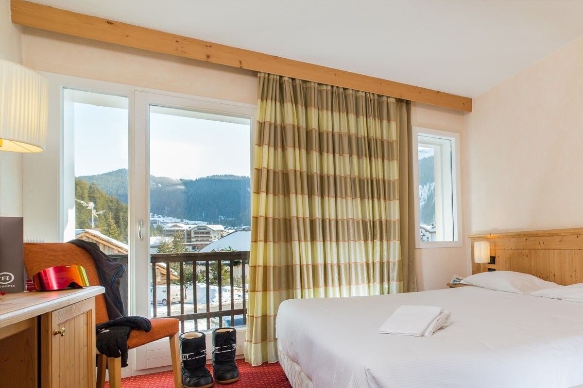 A hotel room with a bed, a desk, a chair, a view of a mountain, and a balcony with a railing.