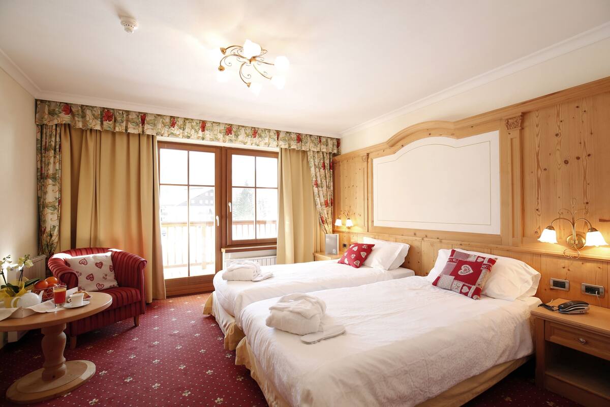 A hotel room with two beds, a sitting area with a red chair, a wooden headboard, and a large window with curtains.