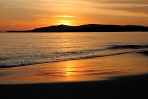Una playa con un atardecer sobre el agua, una orilla arenosa y colinas distantes.