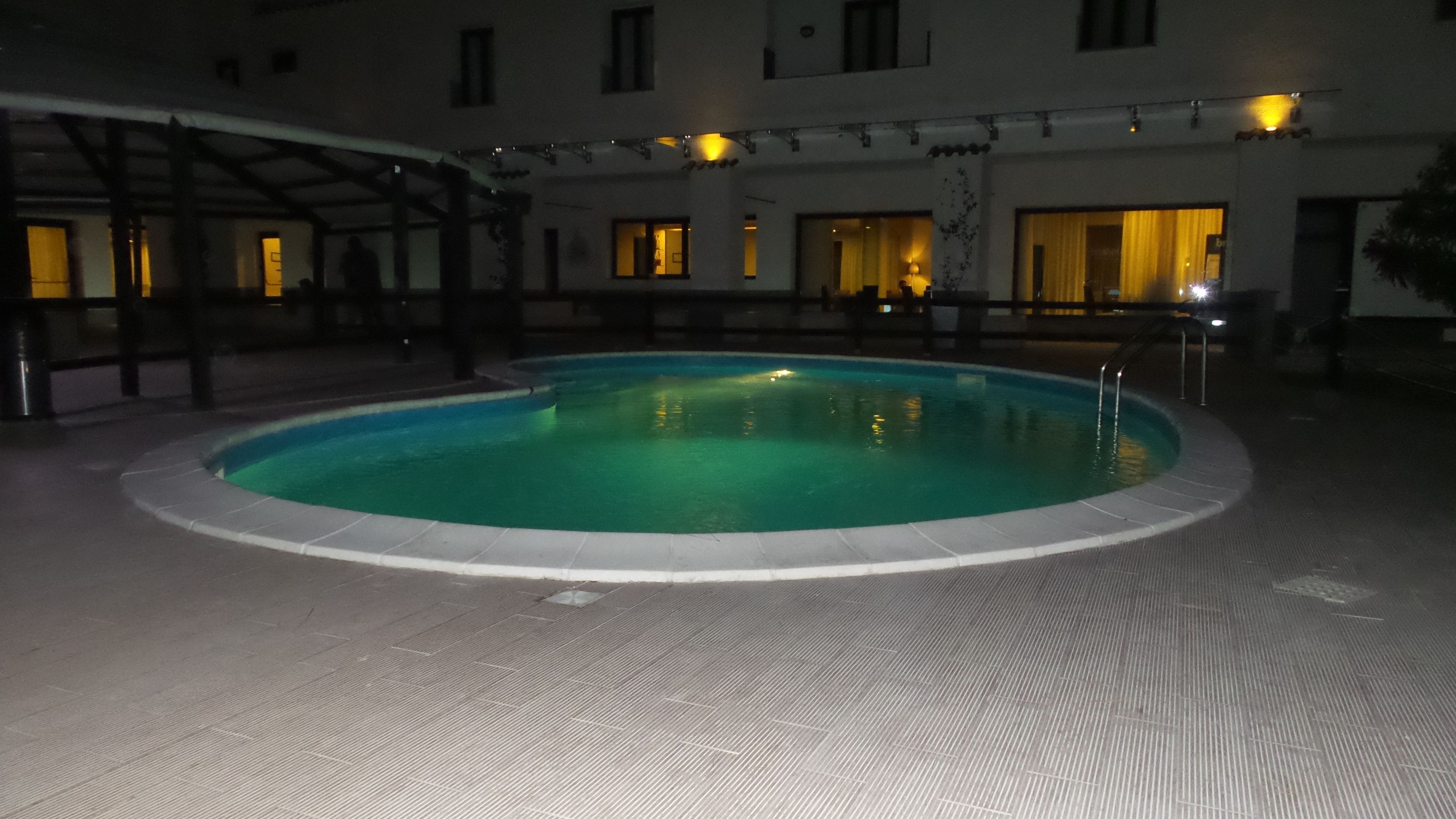 A swimming pool with a tiled deck, surrounded by a building with multiple windows and a covered area.