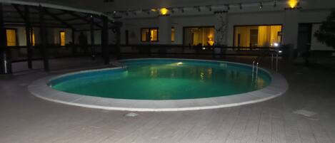 A swimming pool with a tiled deck, surrounded by a building with multiple windows and a covered area.