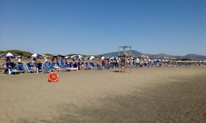 Una spiaggia con file di sedie e ombrelloni azzurri e bianchi, un galleggiante e una torretta di salvataggio.
