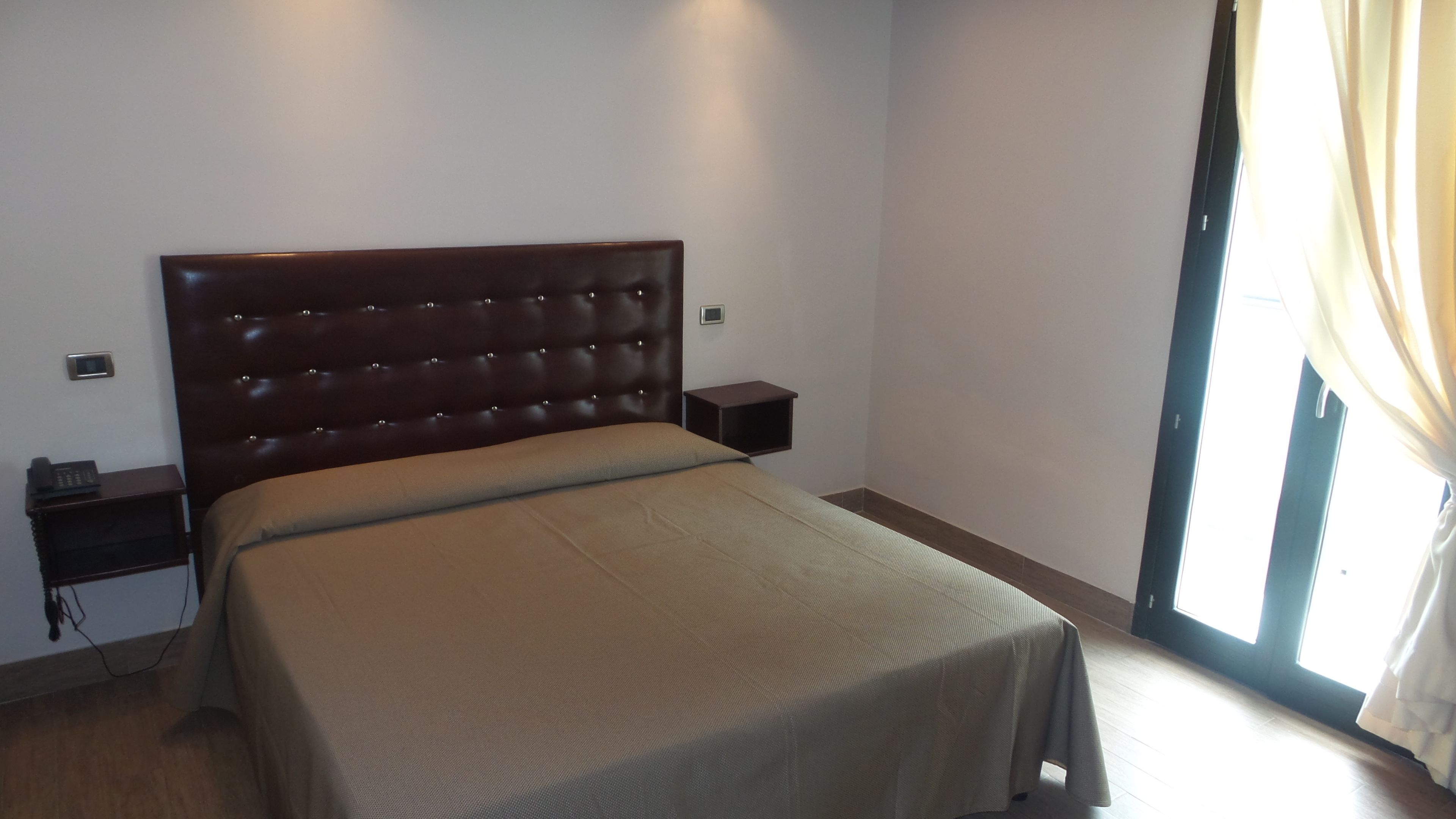 Une chambre à coucher avec un grand lit, une table de chevet, un réveil fixé au mur et une fenêtre avec des rideaux.