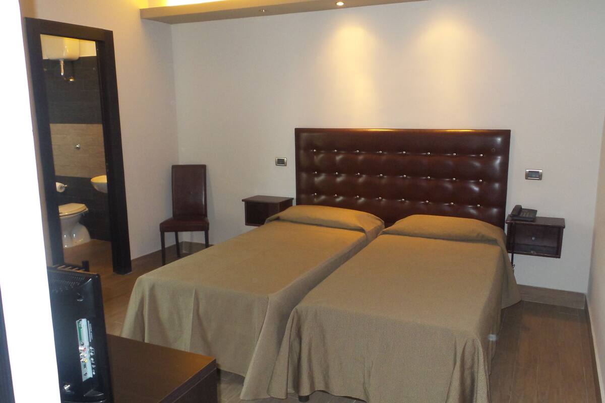 A hotel room with a double bed, a chair, a bedside table, a nightstand, and a bathroom with a toilet and sink.