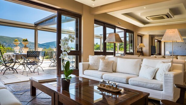 A spacious living area with a white sofa, a wooden coffee table, and a dining area with chairs and a table. Large windows offer a view of the outdoors. - Almyra Hotel (Kefalonia)
