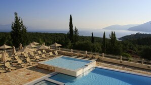 A swimming pool with lounge chairs and umbrellas, surrounded by trees and overlooking a body of water. - Almyra Hotel (Kefalonia)