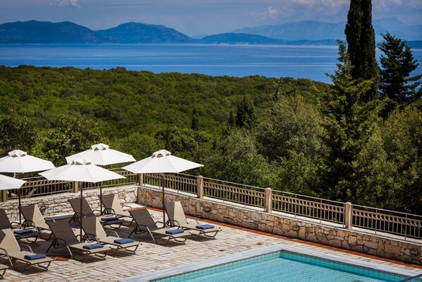 A poolside area with lounge chairs and umbrellas, surrounded by lush greenery and a view of the sea.