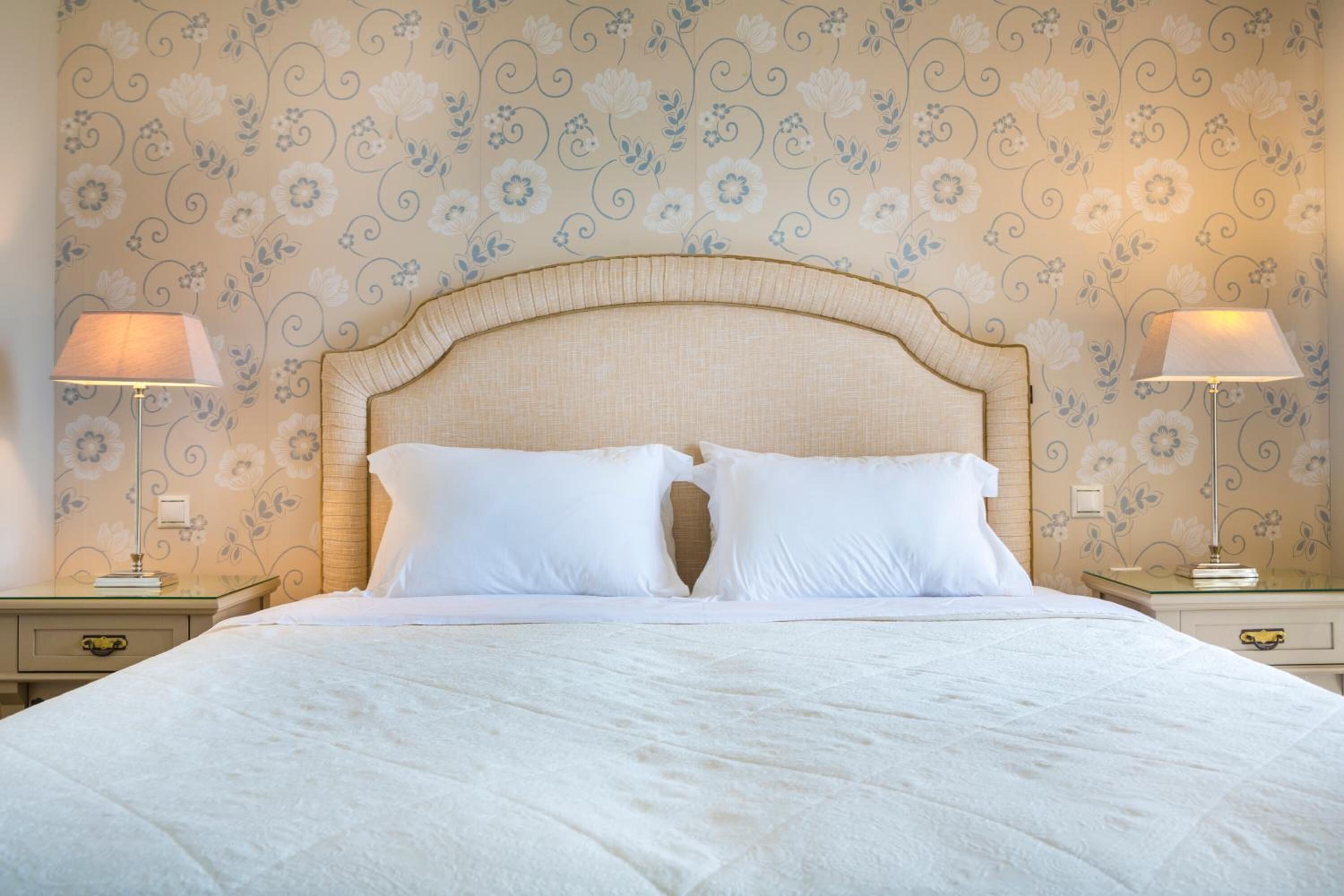 A neatly made bed with a wooden headboard, two bedside lamps, and a floral wallpaper pattern.