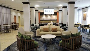 Un hall de hotel con una pantalla grande que muestra el nombre del hotel, un área de descanso con sofás y sillones, y un comedor al fondo.