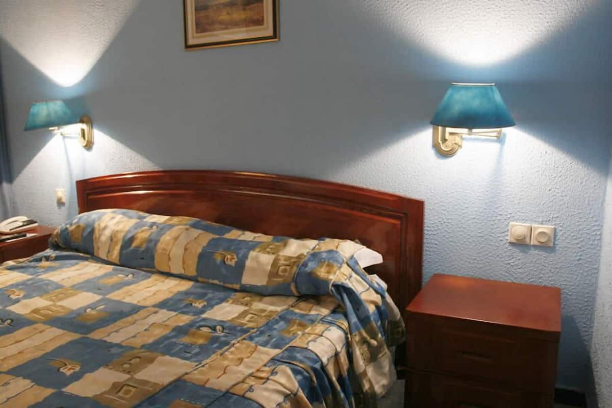 A bedroom with a wooden headboard, a quilted bedspread, a bedside table with a lamp, and a nightstand.