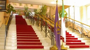 A grand staircase with red carpeted steps and a golden handrail, leading to a landing with a flag and a potted plant. - Safir Mazafran (Algiers)