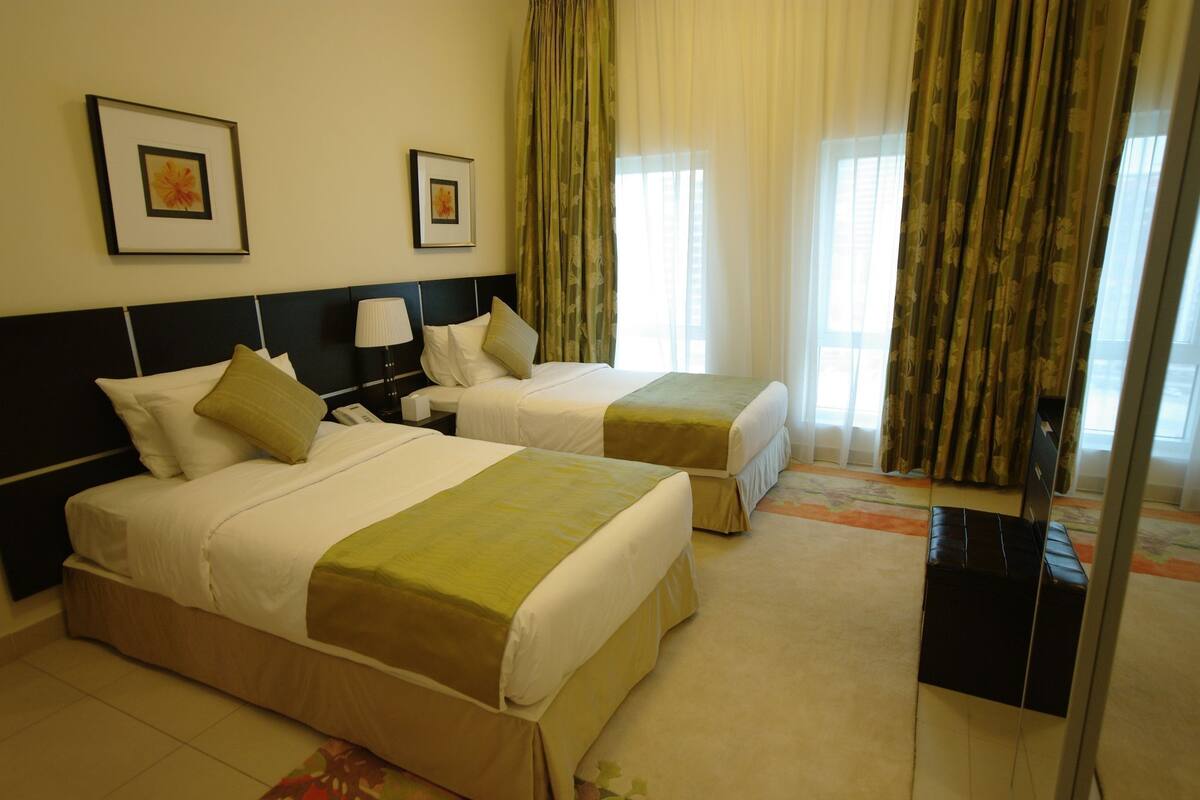 A hotel room with two beds, a nightstand, a lamp, and a view of the outside through a window with curtains.