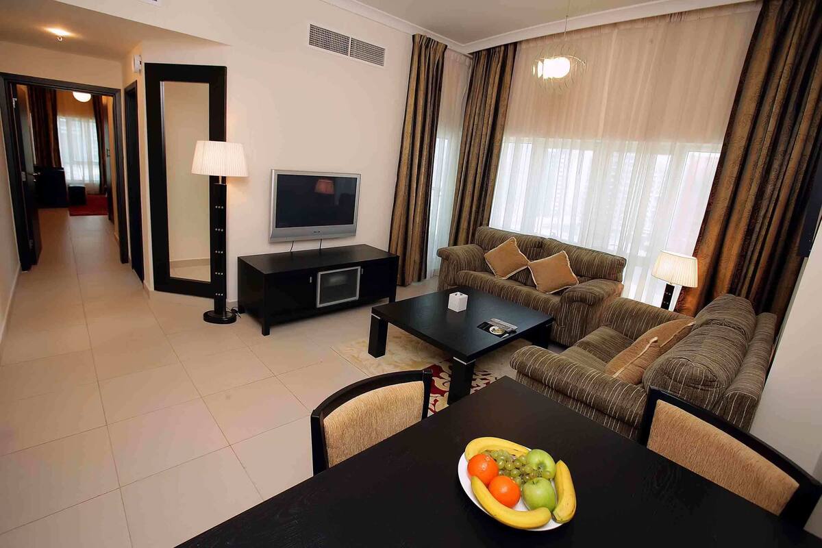 A modern living room with a sofa, a TV, a dining table with fruit, and a hallway leading to another room.
