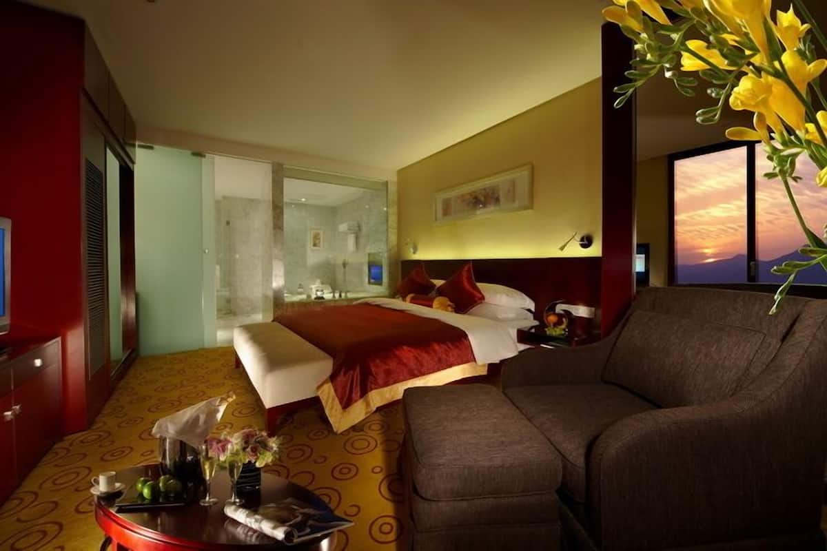 Quarto de hotel com cama, sofá, televisão, banheiro com paredes de vidro e vista para o pôr do sol.