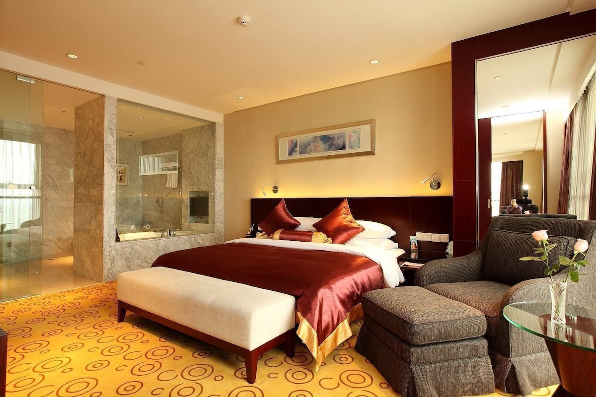 Quarto de hotel com uma cama grande, área de estar com mesa de vidro e banheiro com box de vidro.