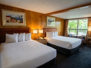 A hotel room with two beds, a desk with a lamp, a telephone, and a window with a view of trees.