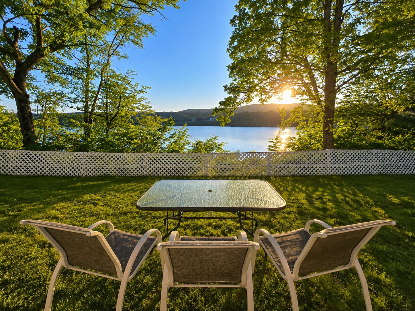 An outdoor seating area with a table and chairs, surrounded by a white fence, overlooking a body of water and hills.