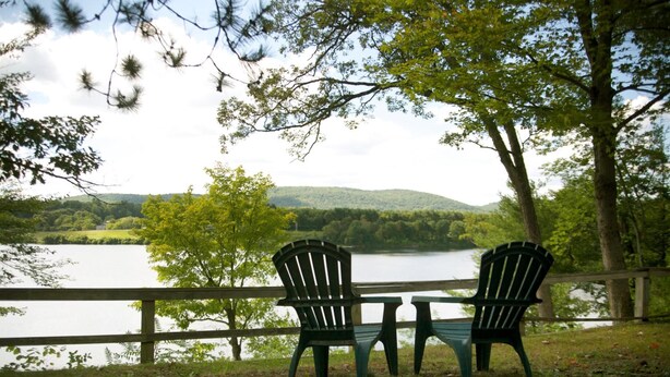 Two Adirondack chairs facing a lake with a wooden deck and a view of a hilly landscape.