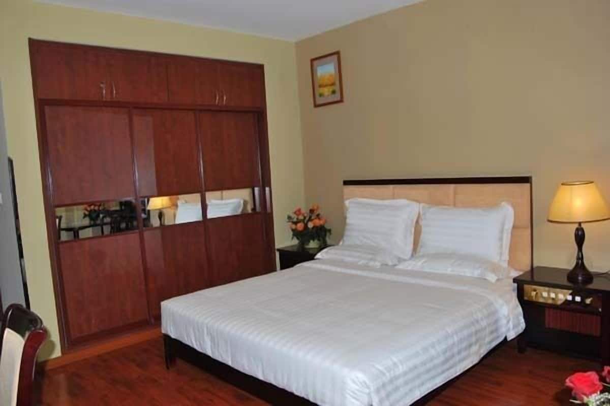 Una habitación de hotel con un armario de madera grande, una cama con ropa blanca, una mesita de noche con lámpara y una pequeña mesa con flores.