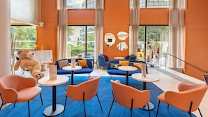 Eine moderne Hotelhalle mit orangefarbenen Wänden, großen Fenstern und einem blauen Teppich. Es gibt orange und blau gefärbte Sessel, einen Teddybären und einen runden Tisch mit einer Speisekarte.