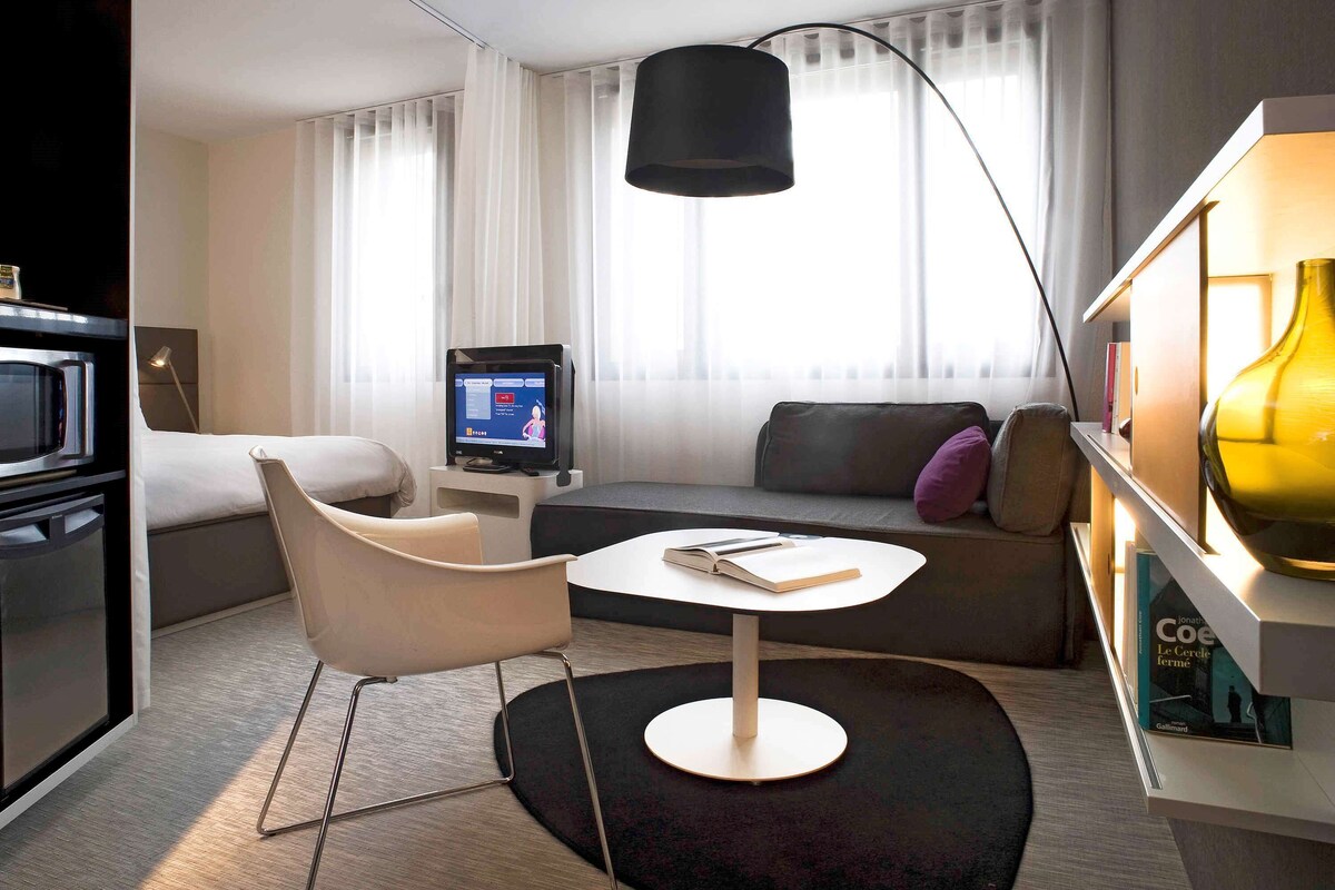 Een moderne hotelkamer met een bank, een klein tafeltje, een tv op een standaard, een lamp en een boekenplank vol boeken.