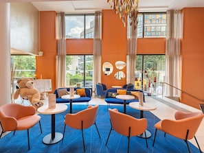 A modern hotel lobby with a blue carpet, orange walls, and large windows. There are sofas, chairs, and round tables. - Novotel Suites Paris Issy les Moulineaux (Issy-les-Moulineaux)