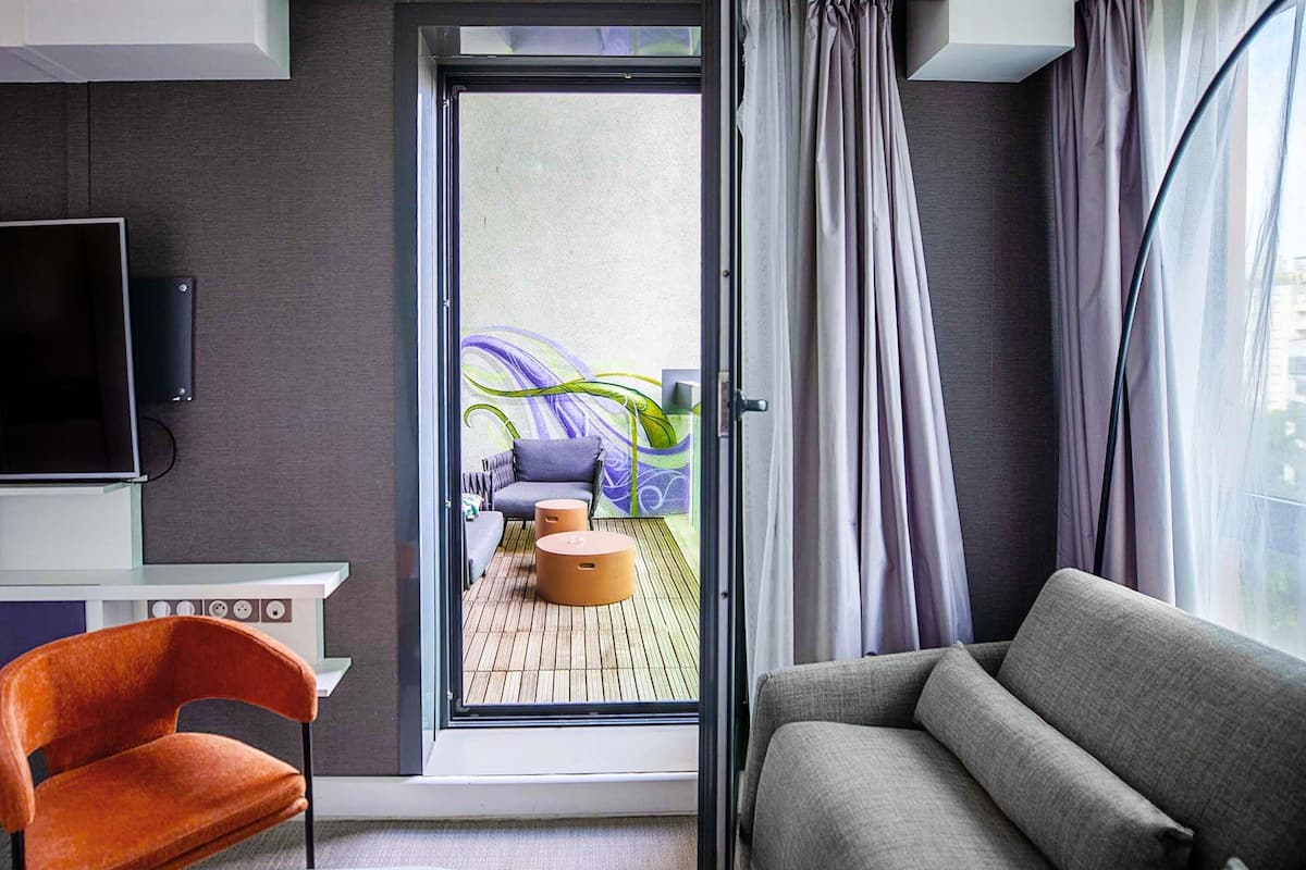 A modern hotel room with a sofa, a chair, a small table, a TV, and a sliding glass door leading to an outdoor area with a couch and a colorful mural.