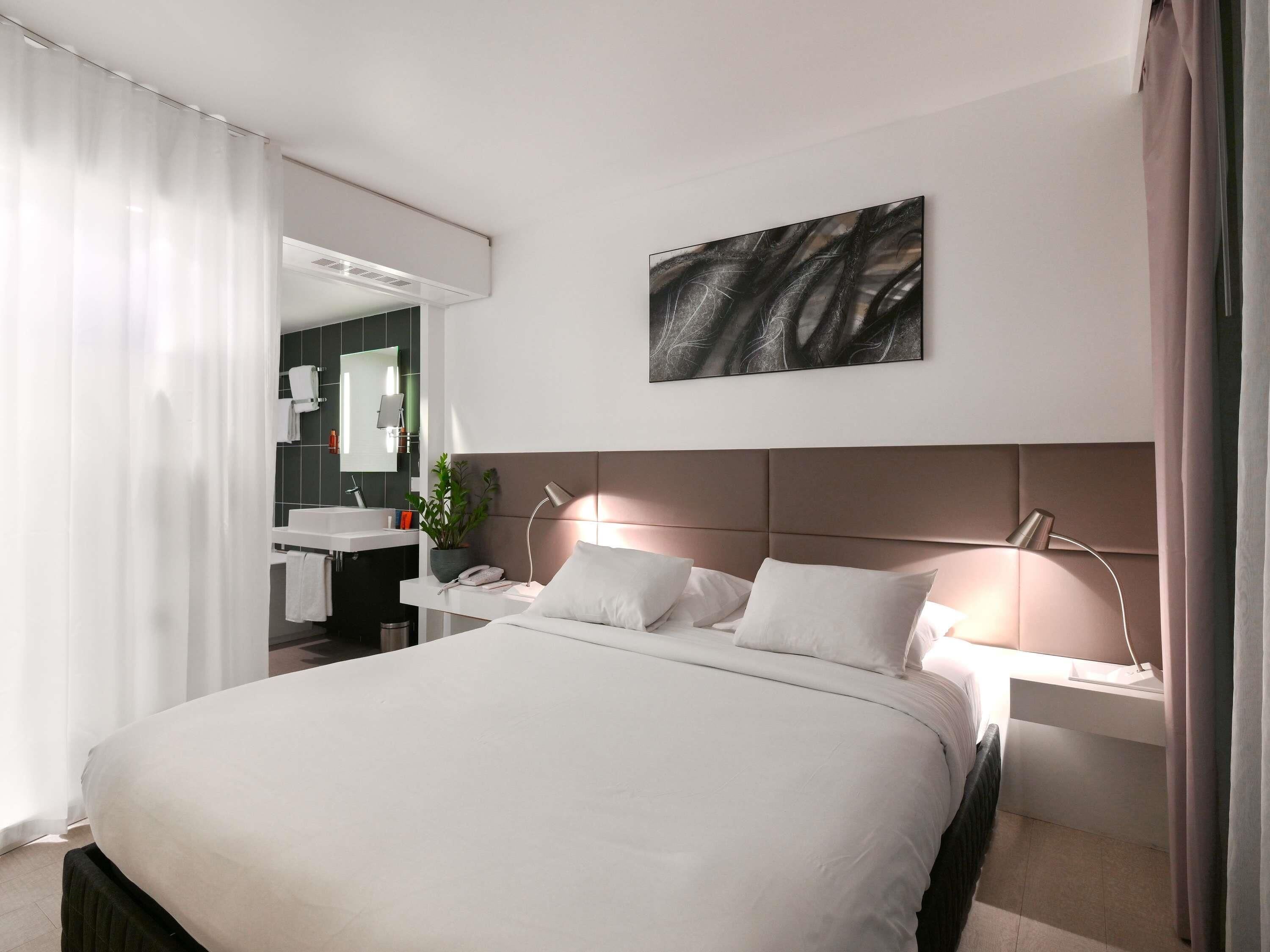 Une chambre d’hôtel moderne avec un grand lit, des tables de chevet, un miroir et une œuvre d’art accrochée au mur.