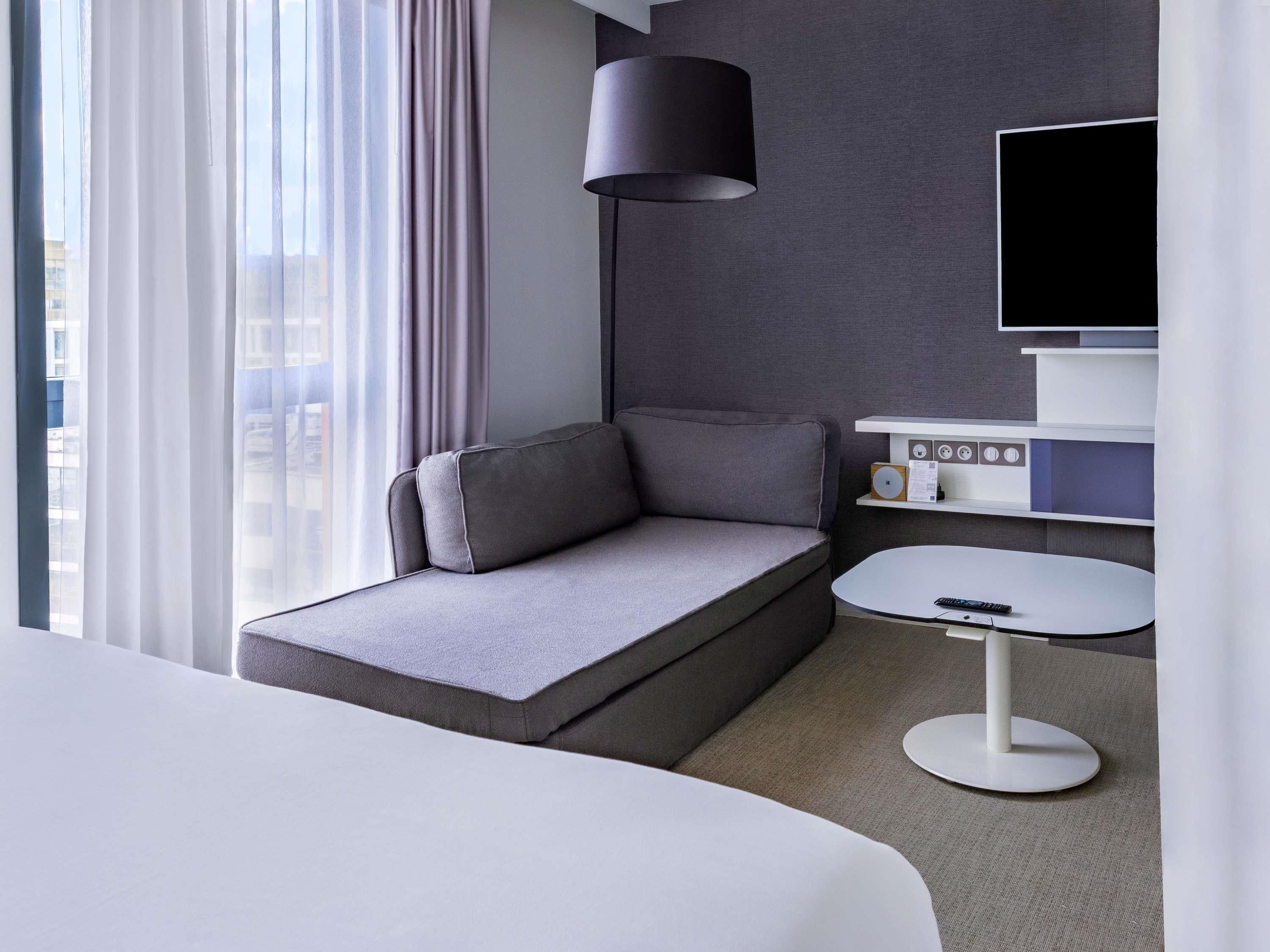 A modern hotel room with a sofa bed, a small round table, a lamp, a TV, and a control panel.