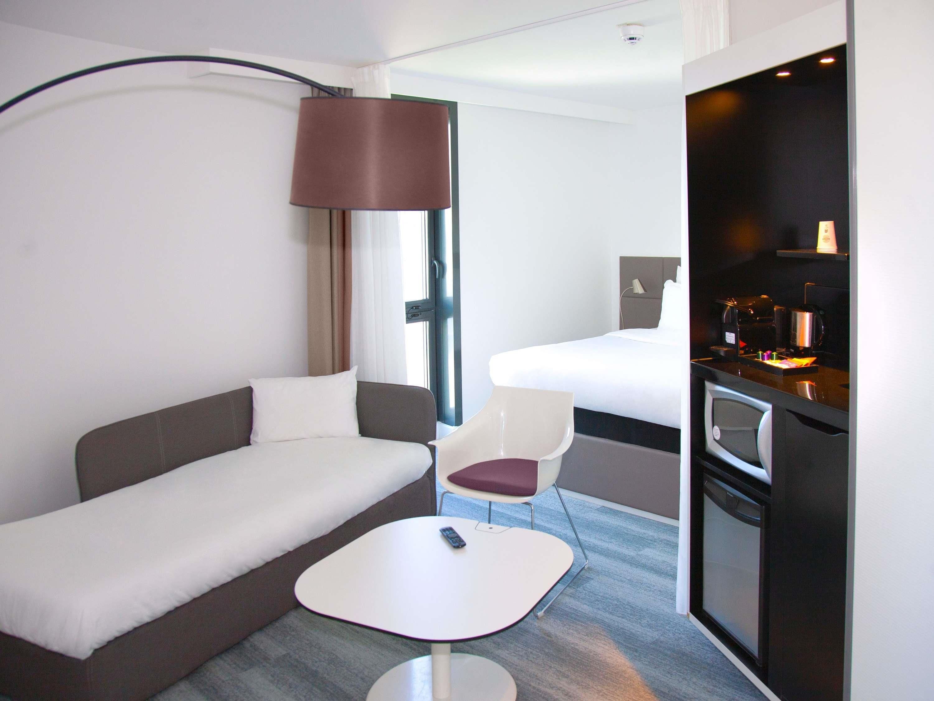 A compact hotel room with a bed, a sofa, a small table, a chair, a kitchenette, and a window.