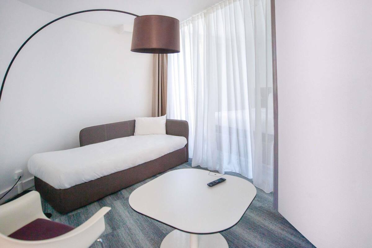 Une chambre d’hôtel avec un lit, une petite table, une chaise, une lampe et une fenêtre avec des rideaux.