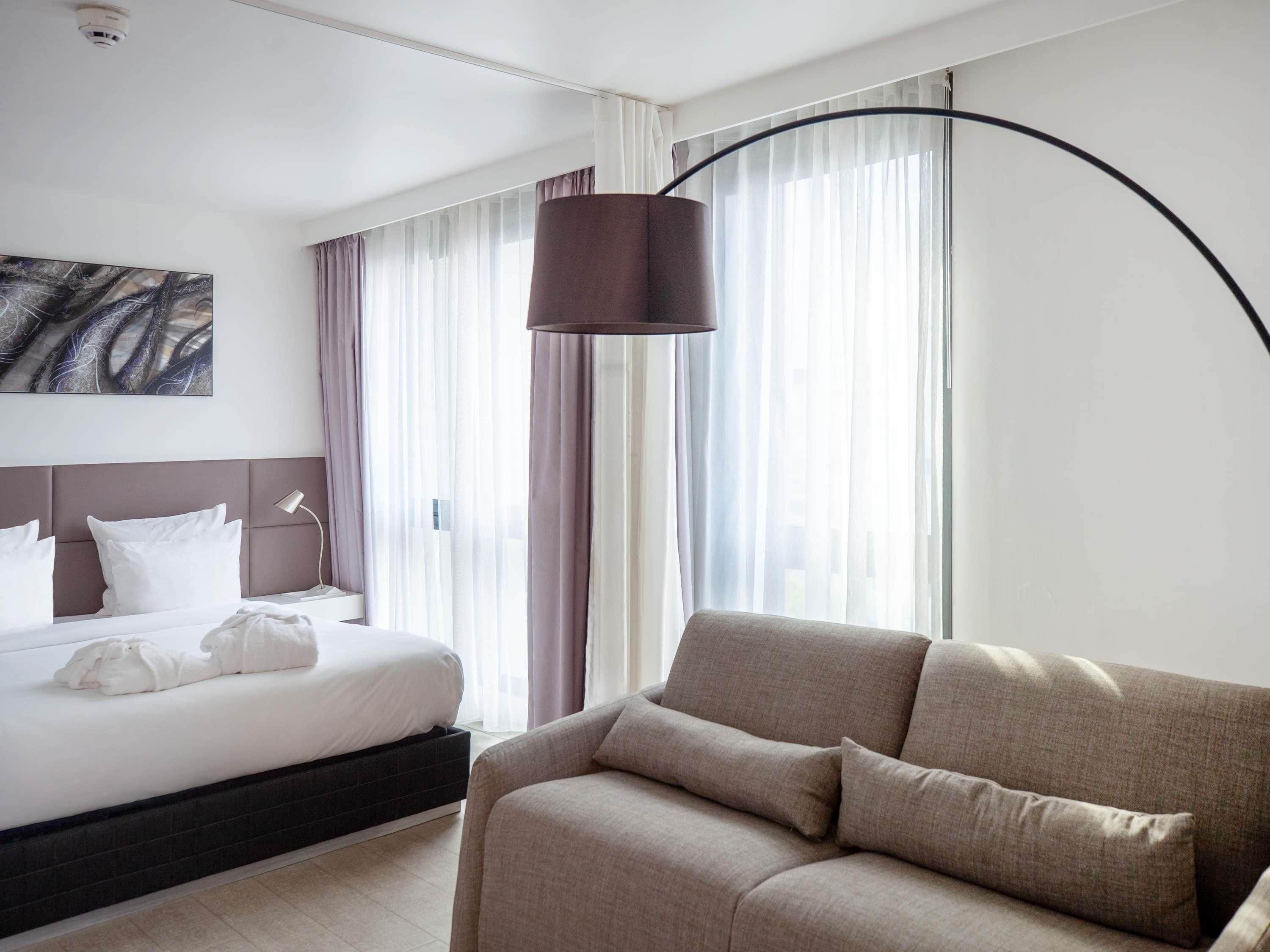 A hotel room with a bed, a sofa, a large window with curtains, a floor lamp, and a painting on the wall.
