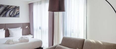 A hotel room with a bed, a sofa, a large window with curtains, a floor lamp, and a painting on the wall.