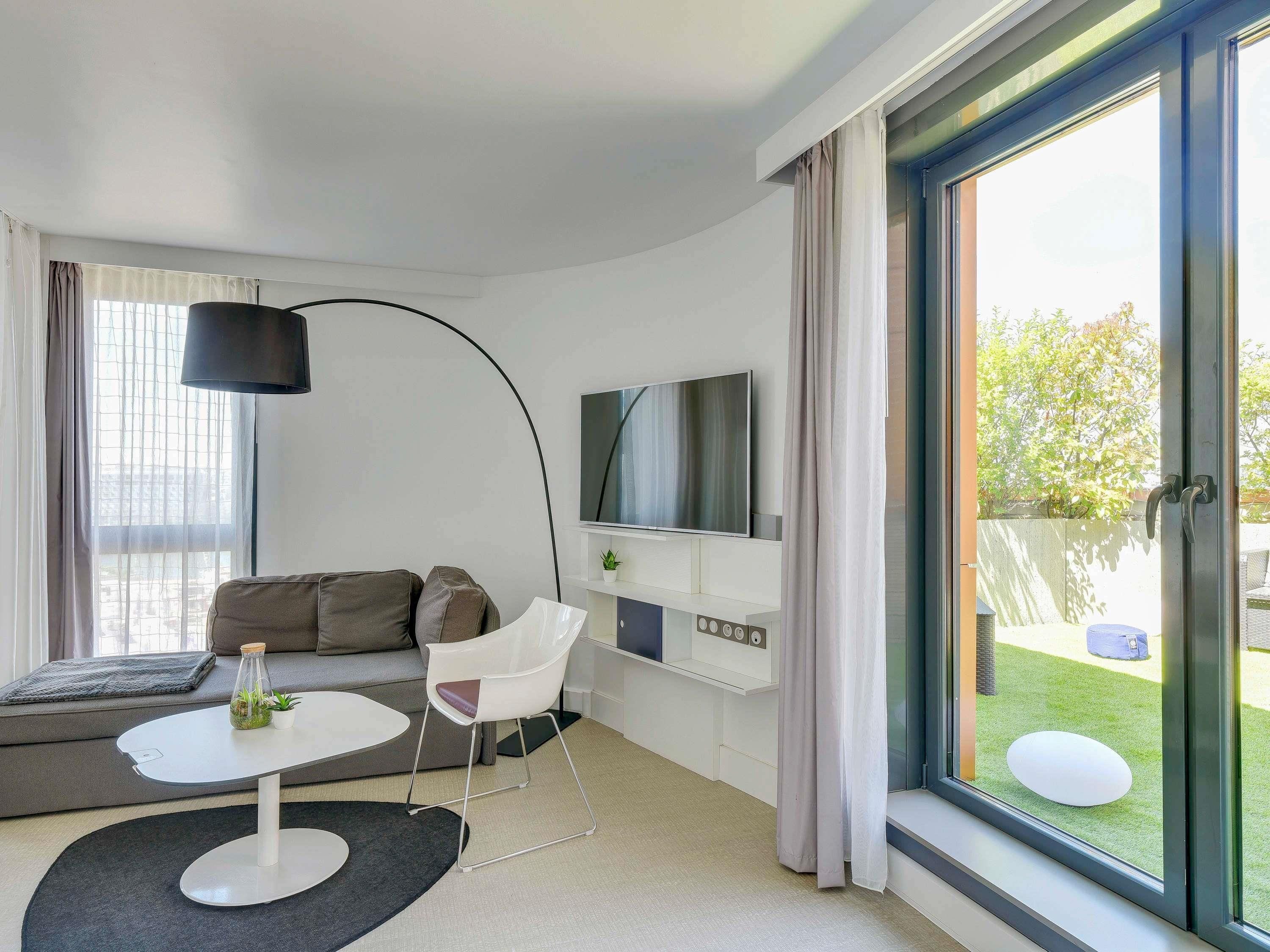 A modern living room with a sofa, a white table, a chair, a TV, and a large window.
