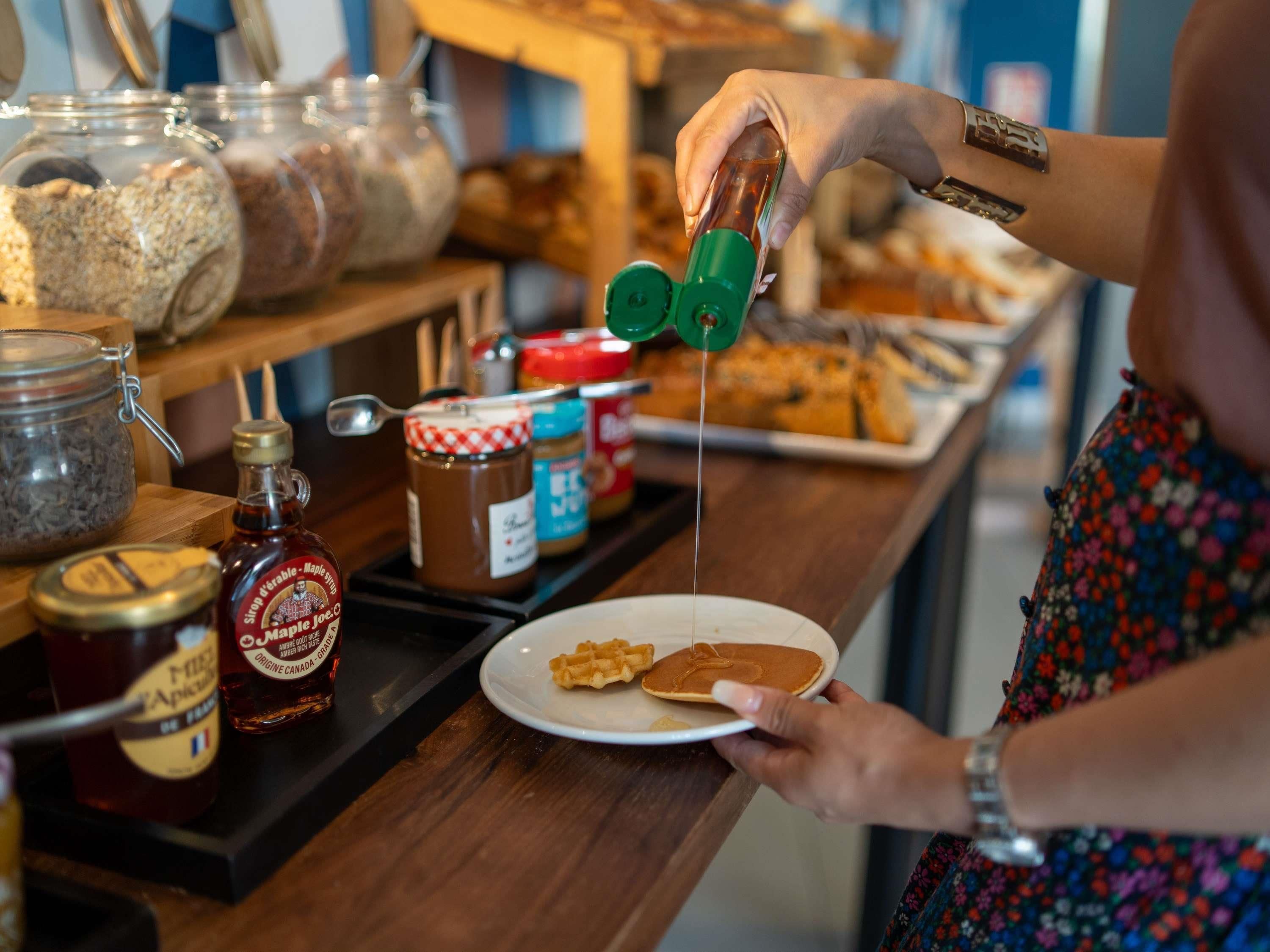 A person pouring maple syrup onto pancakes at a counter with various condiments and food items.
