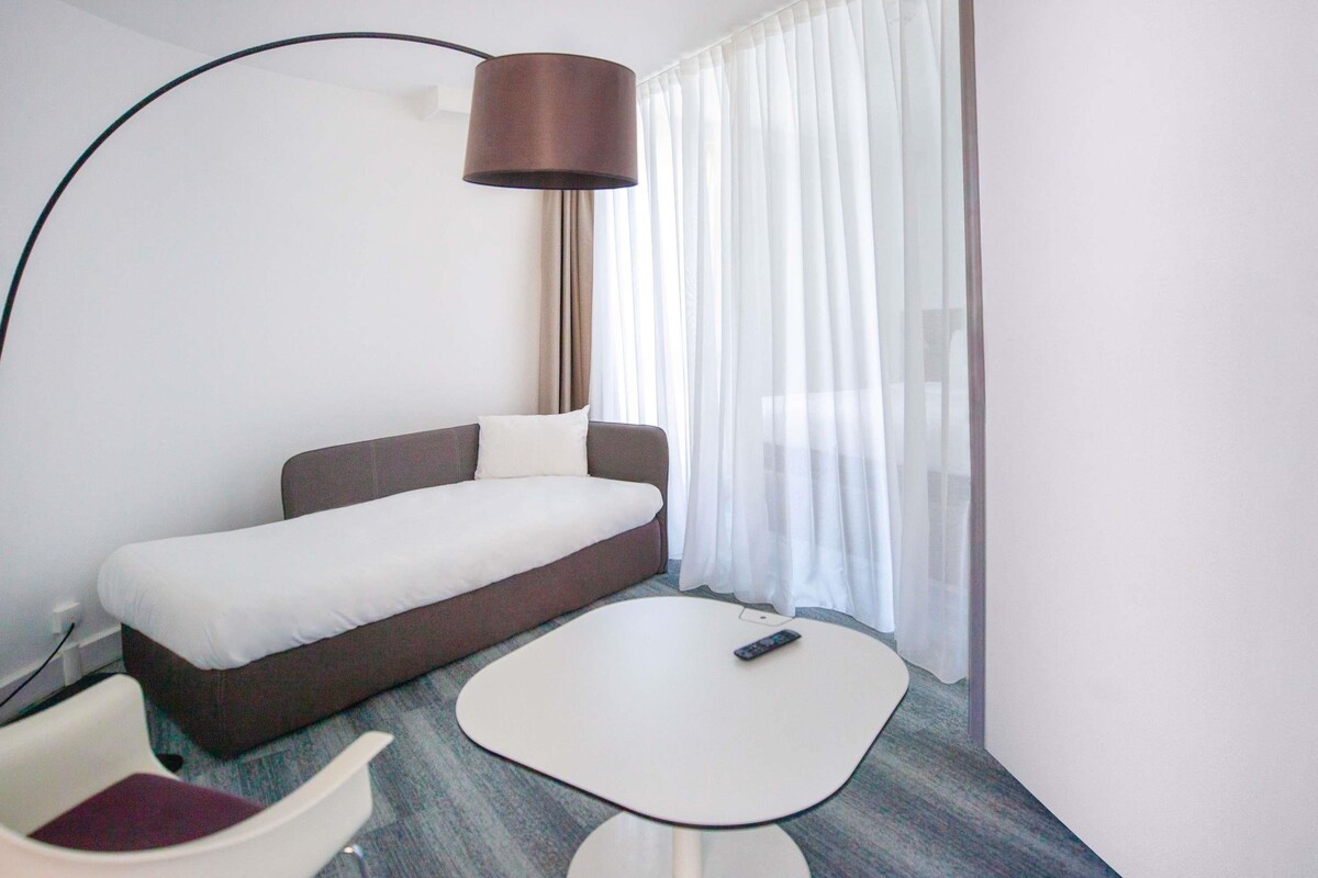 A hotel room with a bed, a small table, a chair, a lamp, and a window with curtains.