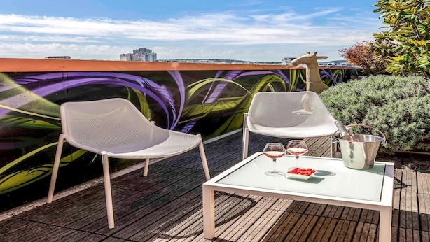 Eine Dachterrasse mit zwei weißen Netzstühlen, einem Glastisch mit einem Weinglas und einem Eimer, sowie einer im Hintergrund sichtbaren Graffiti-Mauer.